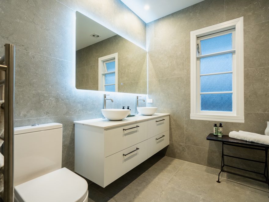Bathroom with grey stone-look tiles, a backlit mirror, white wall-hung vanity and two round basins
