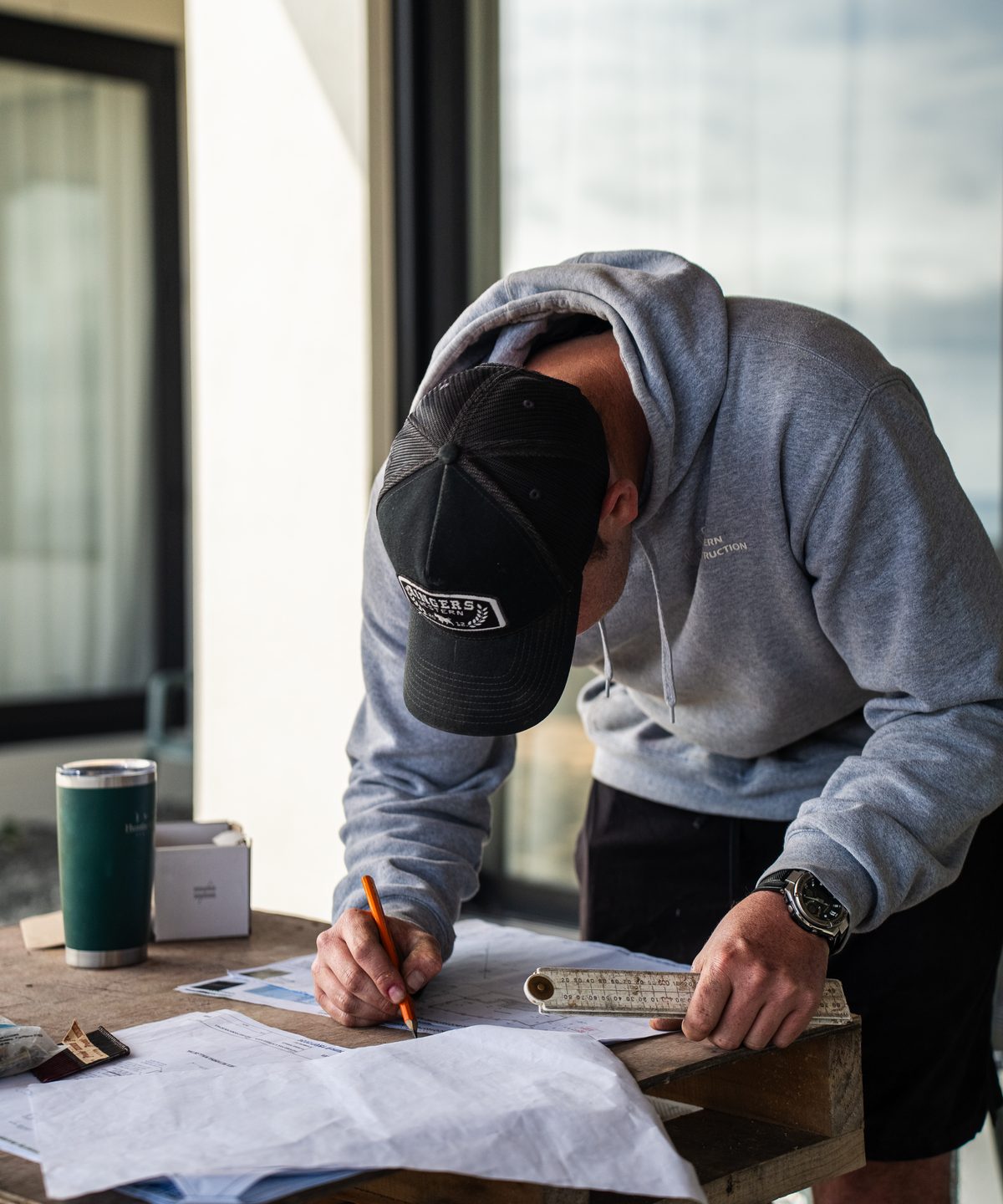 Builder in a cap and hoodie marking up building plans on a table by a window