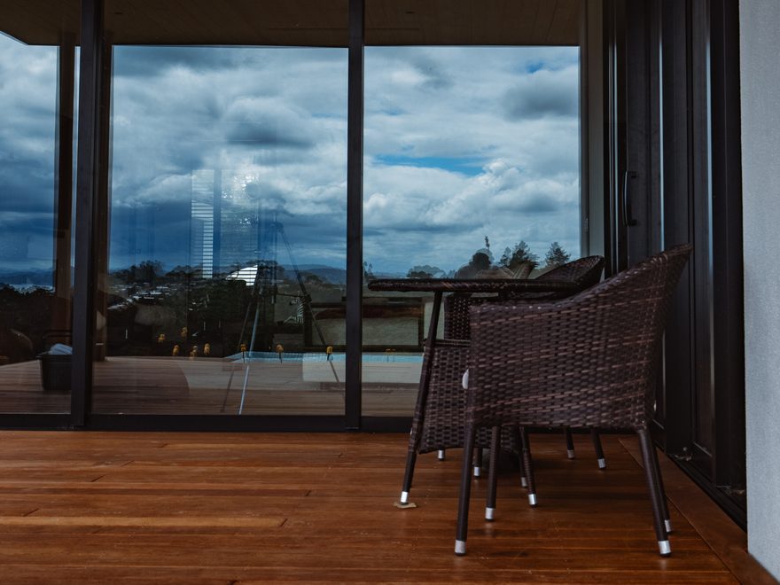 Covered hardwood deck with wicker chairs beside full-height glass doors and a timber-lined ceiling