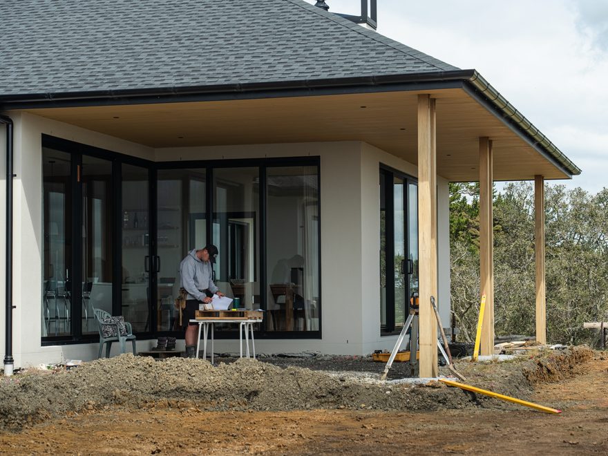 New home with a covered porch on timber posts and a builder working at a table inside the open glass doors